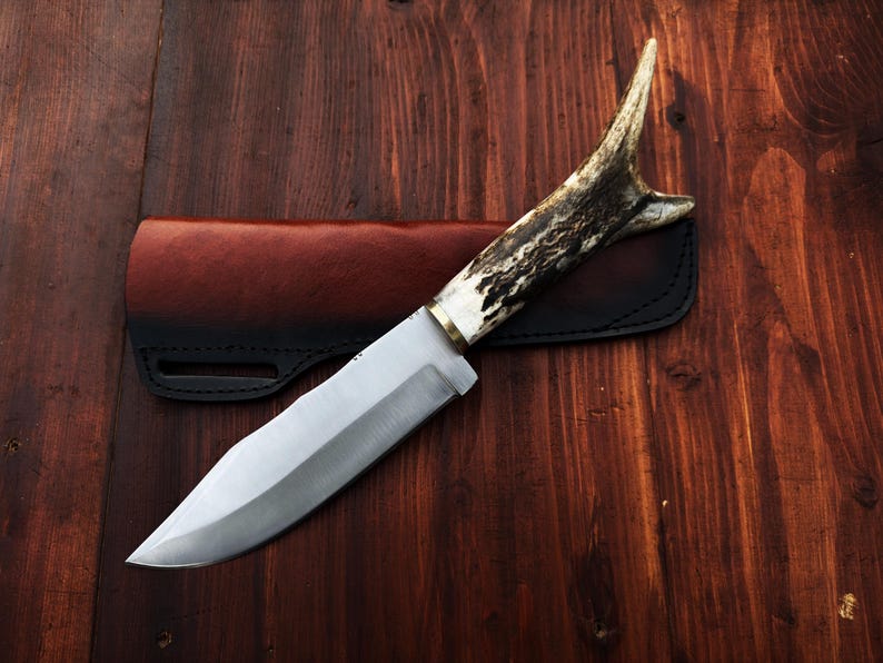 Puede incluir: Cuchillo de caza artesanal con una hoja de acero pulido y un mango de asta. El cuchillo descansa sobre una funda de cuero marr&oacute;n oscuro, sobre un fondo de madera. La hoja mide unos 20 cm.