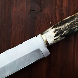 Puede incluir: Primer plano de un cuchillo artesanal con una hoja plateada pulida y un mango de asta y madera. El mango presenta una virola de lat&oacute;n y un patr&oacute;n oscuro y texturizado. El cuchillo descansa sobre una superficie de madera oscura.