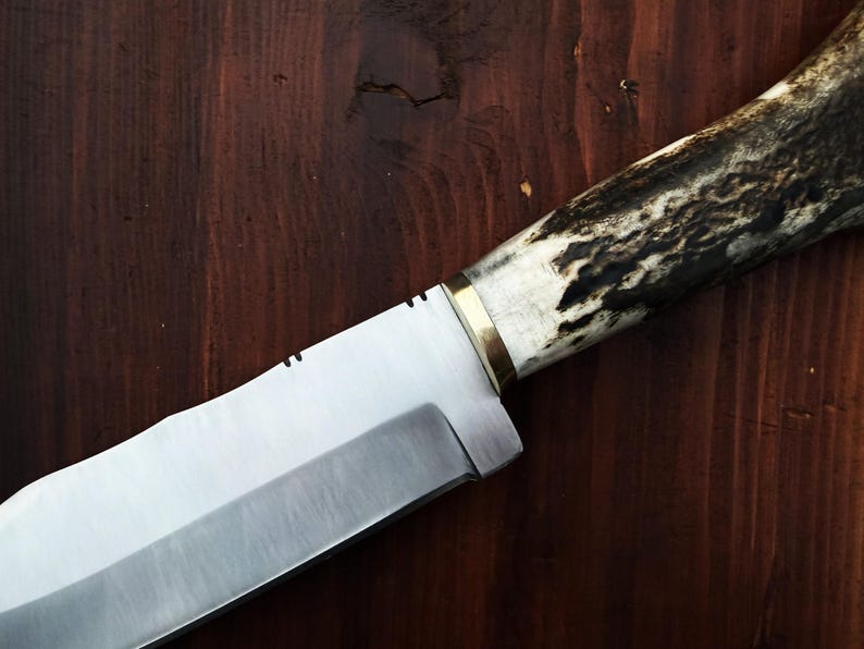 Puede incluir: Primer plano de un cuchillo artesanal con una hoja plateada pulida y un mango de asta y madera. El mango presenta una virola de lat&oacute;n y un patr&oacute;n oscuro y texturizado. El cuchillo descansa sobre una superficie de madera oscura.
