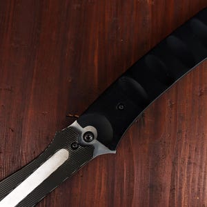 May include: A black and silver knife with a curved blade. The handle is black with a textured design, and the blade has a silver stripe. The knife is set against a dark wood background.