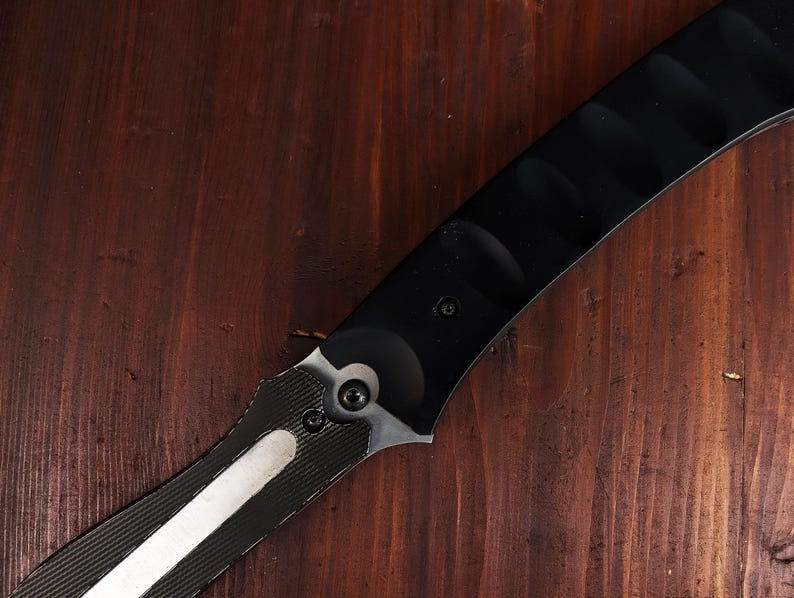 May include: A black and silver knife with a curved blade. The handle is black with a textured design, and the blade has a silver stripe. The knife is set against a dark wood background.