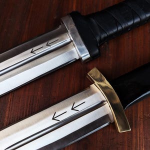 Puede incluir: Dos cuchillos con hojas de metal plateado que presentan dos flechas grabadas apuntando a la izquierda. El cuchillo superior tiene un mango negro con una guarda plateada, mientras que el inferior tiene un mango negro y una guarda de lat&oacute;n.