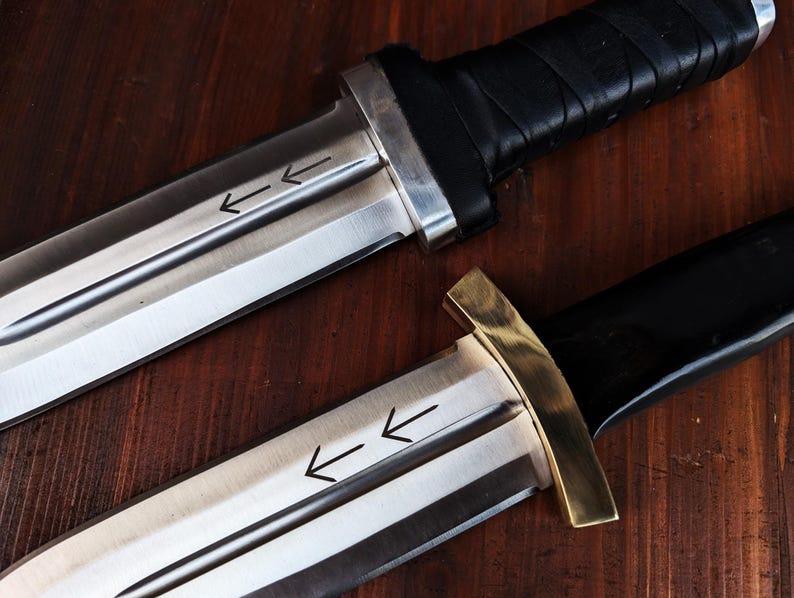Puede incluir: Dos cuchillos con hojas de metal plateado que presentan dos flechas grabadas apuntando a la izquierda. El cuchillo superior tiene un mango negro con una guarda plateada, mientras que el inferior tiene un mango negro y una guarda de lat&oacute;n.