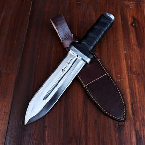 Puede incluir: Un cuchillo de hoja fija con una hoja de acero plateado que presenta un canal central y dos flechas grabadas apuntando hacia la punta. El mango est&aacute; envuelto en material negro y el cuchillo descansa sobre una funda de cuero marr&oacute;n oscuro.