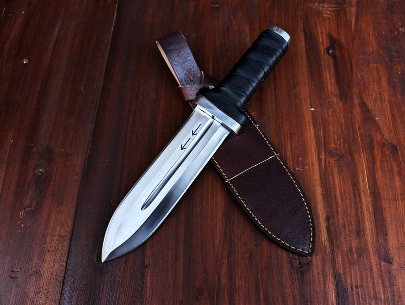 Puede incluir: Un cuchillo de hoja fija con una hoja de acero plateado que presenta un canal central y dos flechas grabadas apuntando hacia la punta. El mango est&aacute; envuelto en material negro y el cuchillo descansa sobre una funda de cuero marr&oacute;n oscuro.