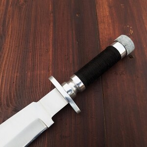 May include: A silver hunting knife with a black handle and a silver pommel. The knife is lying on a wooden surface.