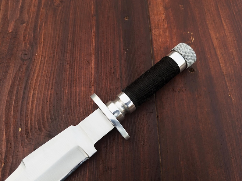 May include: A silver hunting knife with a black handle and a silver pommel. The knife is lying on a wooden surface.
