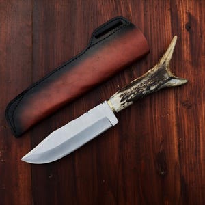 Puede incluir: Un cuchillo de caza con una hoja de acero pulido y un mango de asta. El cuchillo est&aacute; sobre una superficie de madera junto a una funda de cuero con un mango negro y un cuerpo marr&oacute;n rojizo.