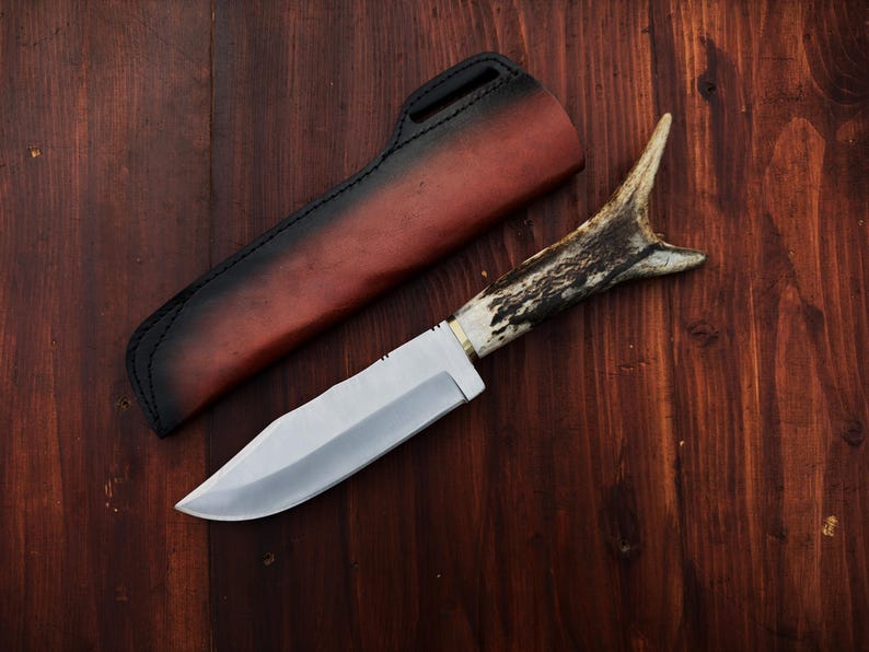 Puede incluir: Un cuchillo de caza con una hoja de acero pulido y un mango de asta. El cuchillo est&aacute; sobre una superficie de madera junto a una funda de cuero con un mango negro y un cuerpo marr&oacute;n rojizo.