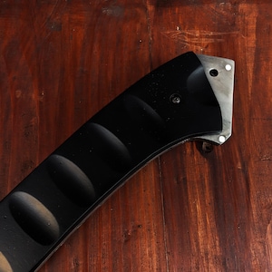 May include: Close-up of a black knife handle with a textured grip and a silver blade. The handle has a curved design with rounded indentations for a secure hold. The blade has a hole and two rivets.