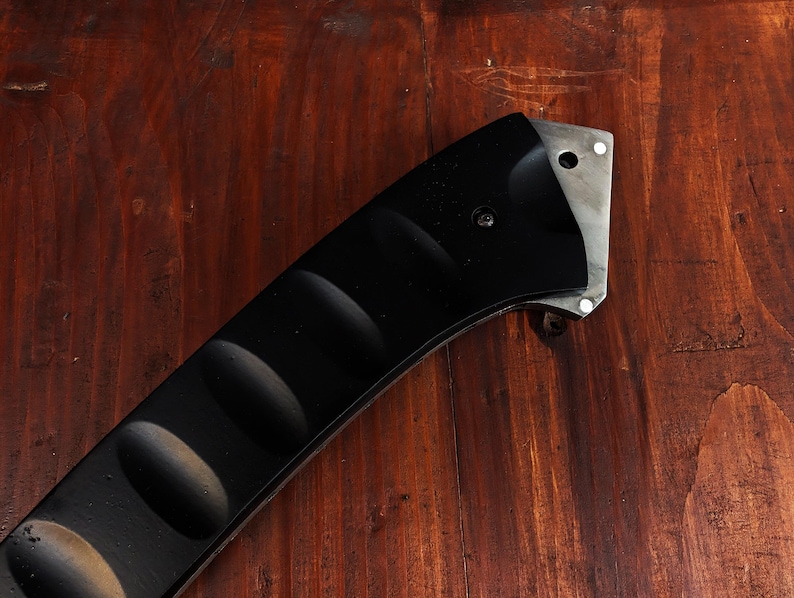 May include: Close-up of a black knife handle with a textured grip and a silver blade. The handle has a curved design with rounded indentations for a secure hold. The blade has a hole and two rivets.