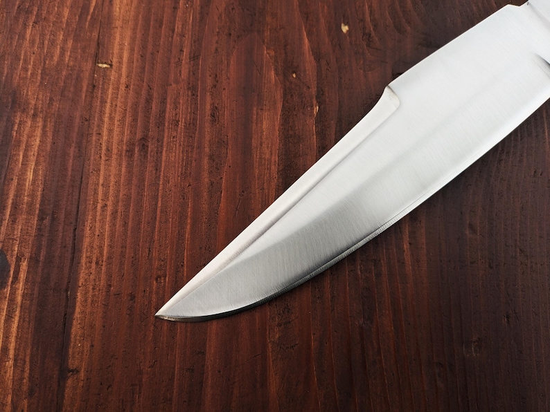 May include: A close-up of a silver knife blade on a dark brown wooden surface.