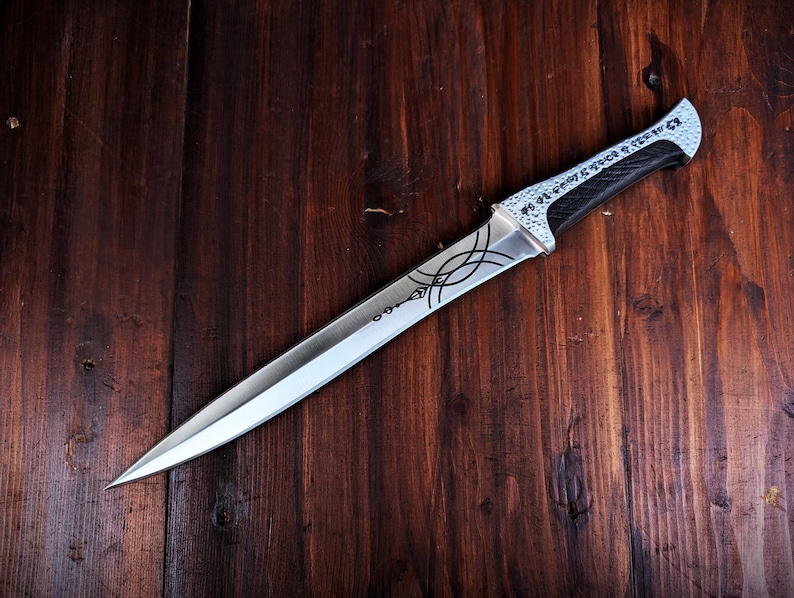 May include: A silver dagger with a pointed blade and a black handle. The blade features etched designs and the handle has a textured grip. The dagger rests on a dark wooden surface.