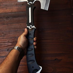May include: A tactical axe with a black and silver design. The axe head features a cutting blade, a hammer, and a spike. The handle is black with a textured grip. The axe is held against a dark wood background.