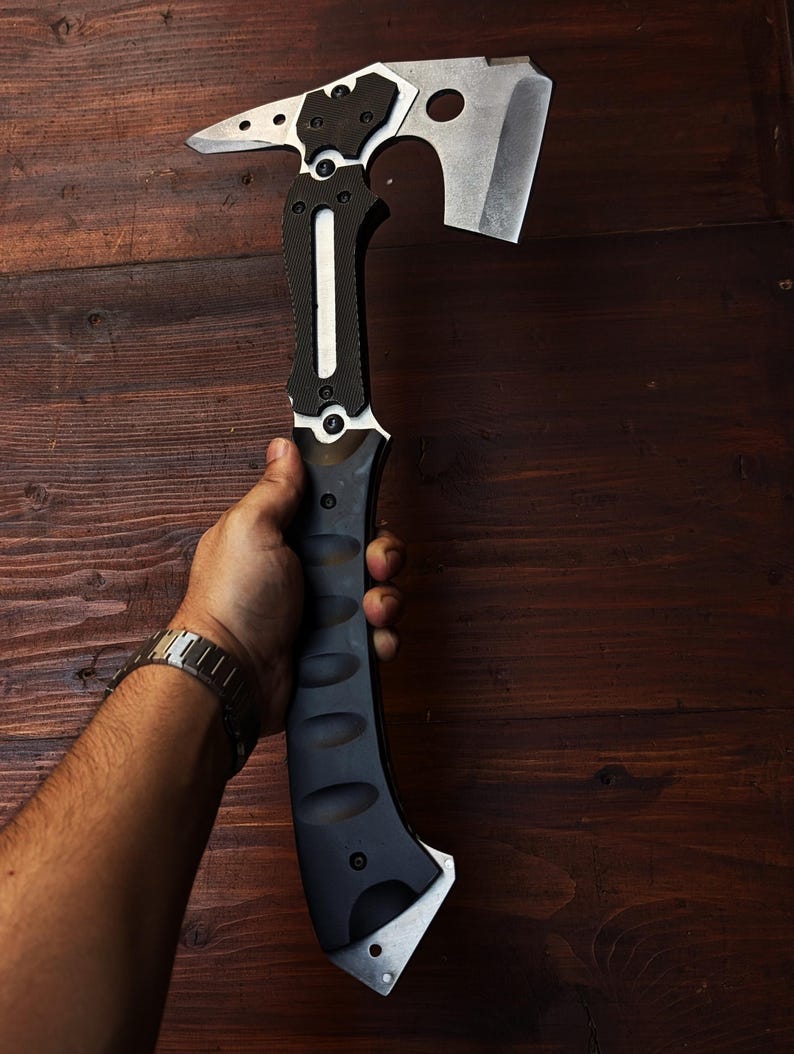 May include: A tactical axe with a black and silver design. The axe head features a cutting blade, a hammer, and a spike. The handle is black with a textured grip. The axe is held against a dark wood background.