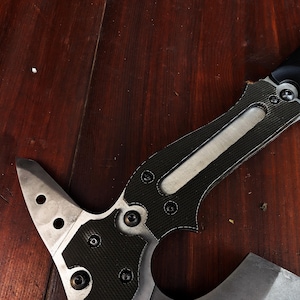 May include: A tactical axe with a black and silver blade and handle. The handle features a carbon fibre pattern and is secured with multiple screws. The axe head has a sharp edge and several circular cutouts.