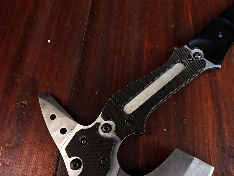 May include: A tactical axe with a black and silver blade and handle. The handle features a carbon fibre pattern and is secured with multiple screws. The axe head has a sharp edge and several circular cutouts.