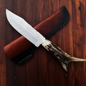 Puede incluir: Un cuchillo de caza con una hoja plateada y un mango de asta. El cuchillo est&aacute; sobre una funda de cuero marr&oacute;n con borde negro, sobre un fondo de madera oscura. La hoja del cuchillo mide unos 20 cm.