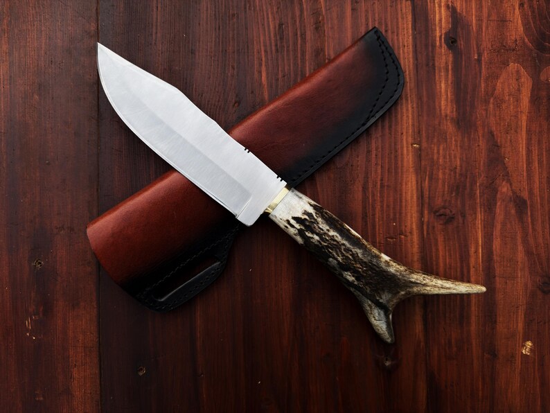 Puede incluir: Un cuchillo de caza con una hoja plateada y un mango de asta. El cuchillo est&aacute; sobre una funda de cuero marr&oacute;n con borde negro, sobre un fondo de madera oscura. La hoja del cuchillo mide unos 20 cm.