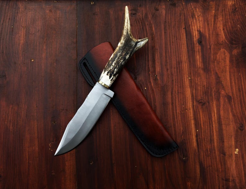 Puede incluir: Un cuchillo de caza con una hoja de acero pulido y un mango de asta. El cuchillo descansa sobre una funda de cuero marr&oacute;n con costuras negras. El cuchillo y la funda se colocan sobre un fondo de madera oscura.