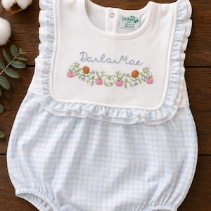 May include: A white and light blue gingham bubble romper featuring a white bib front with ruffled edges. The bib is embroidered with the name Darla Mae in blue script above a vine pattern with small pink and orange pumpkins and tiny flowers.