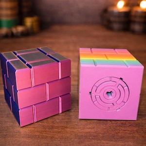 May include: Two colorful puzzle cubes. One is pink with a rainbow top and a circular maze design. The other is pink and blue, with a brick-like pattern. The cubes are on a wooden surface.