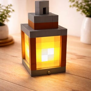 May include: A gray and orange block-style lantern with a bright yellow light emanating from within. The lantern is made of square blocks and sits on a wooden surface. The light is on.