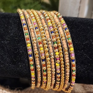 May include: A set of 10 gold bangles with colorful beads. The bangles are stacked on top of each other and have a wooden base.