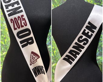 May include: White satin sash with black glitter text reading "Senior 2025" and "Kylie Hansen". The sash has a maroon cheerleading megaphone with the text "Cheer" and "WWHS" on it.