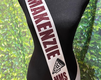 May include: White satin sash with maroon glitter text that reads "2025 McKenzie Cheer MMS Captain". The sash is draped over a black mannequin. A megaphone graphic is also included.