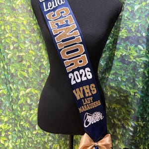 May include: A navy blue sash featuring the text "CAPTAIN Leila", "SENIOR", "2026", "WHS", "LADY MARAUDERS", and "Cheer" in white and gold glitter lettering. The sash is displayed on a black mannequin and includes a gold satin bow at the bottom.