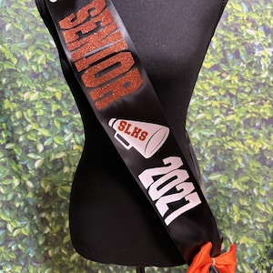 May include: A black satin sash featuring the name Georgia in white script, the word SENIOR in orange glitter, a megaphone graphic with the text SLHS, the year 2021 in white, and an orange ribbon bow attached at the bottom end of the sash.