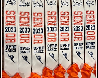 May include: Eight white satin sashes with orange bows. Each sash has a name, "Senior 2023", and "OPRF Dance" printed on it. The sashes are arranged in a row with the bows facing the viewer. The text "Not Without A Bow™" is printed at the bottom of the image.