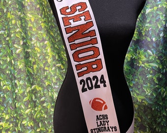 May include: A white satin sash featuring the name Saige in black script, the word SENIOR in orange glitter, the year 2024 in black, an orange football graphic, and the text ACHS LADY STINGRAYS in black, finished with a black satin bow at the bottom.