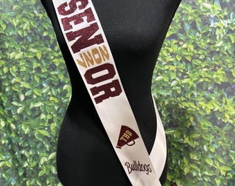 May include: A white satin sash featuring glittery maroon and gold text that reads CO CAPTAIN Audrie SENIOR 2027, a maroon megaphone icon with THS, and the word Bulldogs. The sash is finished with a maroon satin bow at the bottom hip area.