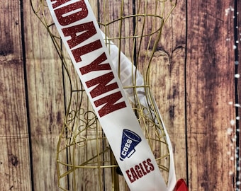 May include: A white satin sash featuring the year 2026 in dark blue, the name ADALYNN in red glitter, a blue megaphone graphic with the text CGES, the word EAGLES in red glitter, and a red satin bow at the bottom, displayed on a gold wire mannequin.