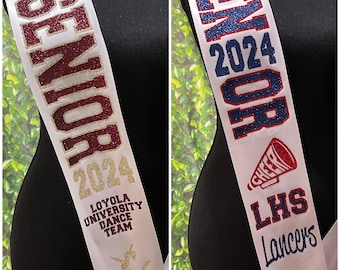 Può includere: Due fasce di raso bianco con testo glitterato. Una fascia recita "Captain Chloe SENIOR 2024 Loyola University Dance Team". L'altra fascia recita "Cadance SENIOR 2024 LHS Lancers Cheer". Entrambe hanno fiocchi.