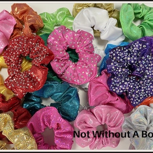 May include: A collection of assorted fabric hair scrunchies in various colors including pink, red, purple, gold, green, blue, white, and silver. Several scrunchies feature a metallic finish or embedded rhinestones. The text Not Without A Bow is visible.