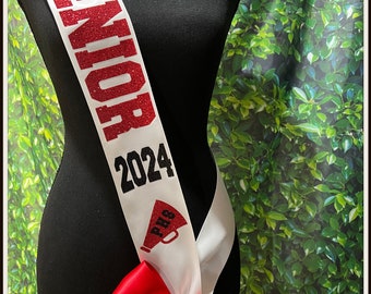 May include: White graduation sash with red glitter lettering that says "Senior 2024" and a red glitter megaphone with the letters "PHS". The sash is tied with a red bow. The text "Not Without A Bow™" is at the bottom of the image.