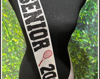 May include: White satin sash with black glitter text that reads "Senior 2023 HHS" and a red tennis racket graphic. The sash also has the name "Valeria" in red text. The text "Not Without A Bow" is printed in red at the bottom of the sash.