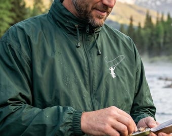 May include: A dark green, long-sleeved, zip-up windbreaker jacket featuring a high collar with drawstring toggles and a small white graphic of a fishing rod and fish on the left chest, worn over light-colored trousers with a fishing net visible.