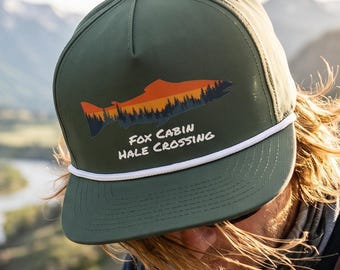 May include: An olive green baseball cap with a white rope detail across the brim. The front features a graphic of a fish silhouette filled with an orange sunset and dark forest, with the text Fox Cabin Hale Crossing printed in white below.