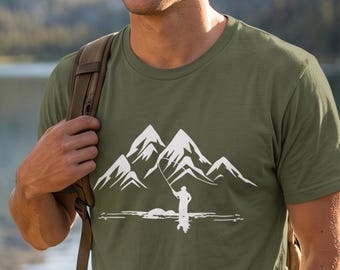 May include: An olive green short-sleeved t-shirt featuring a white graphic print of a mountain range and a person fishing by a lake. The shirt has a crew neckline and is worn with a white baseball cap and a brown backpack with shoulder straps.