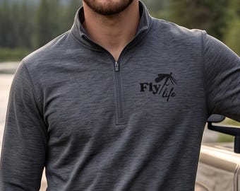 May include: A charcoal gray long-sleeve quarter-zip pullover featuring a black graphic on the left chest that reads Fly Life with a small fishing lure icon. The garment has a textured heathered fabric finish and a stand-up collar with a zipper.