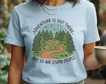 May include: A light blue t-shirt featuring a graphic illustration of a winding path through a forest with pine trees, stars, and a crescent moon. The text reads, "ADVENTURE IS OUT THERE. BUT SO ARE STUPID PEOPLE" in a circular black font.