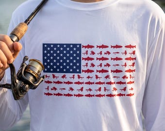 May include: A white long-sleeve shirt featuring a graphic of the American flag. The blue canton contains white stars, while the stripes are composed of repeating red silhouettes of various fish and fishing lures arranged in horizontal rows.
