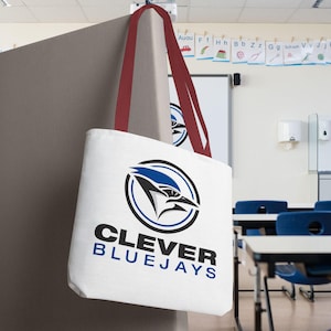 Op de afbeelding: Witte draagtas met bordeauxrode bandjes, met een blauwe gaai-logo in een zwarte cirkel. De tekst "CLEVER BLUEJAYS" staat eronder in zwart en blauw. De tas hangt in een klaslokaal.