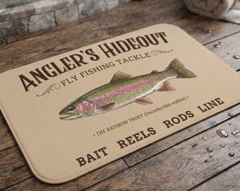 May include: A rectangular tan floor mat featuring the dark brown text ANGLER'S HIDEOUT FLY FISHING TACKLE and BAIT REELS RODS LINE. A detailed illustration of a rainbow trout is centered with the text THE RAINBOW TROUT (Oncorhynchus mykiss).