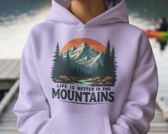 Può includere: Felpa con cappuccio lilla chiaro con grafica di montagna. La grafica presenta una catena montuosa, alberi e la scritta "LIFE IS BETTER IN THE MOUNTAINS". La felpa ha una tasca frontale e un cappuccio.