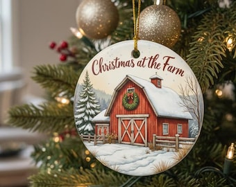 May include: A round Christmas ornament with a winter farm scene. The ornament features a red barn with snow-covered roof and a wreath. The text "Christmas at the Farm" is written on the top. The ornament hangs from a gold string on a Christmas tree.
